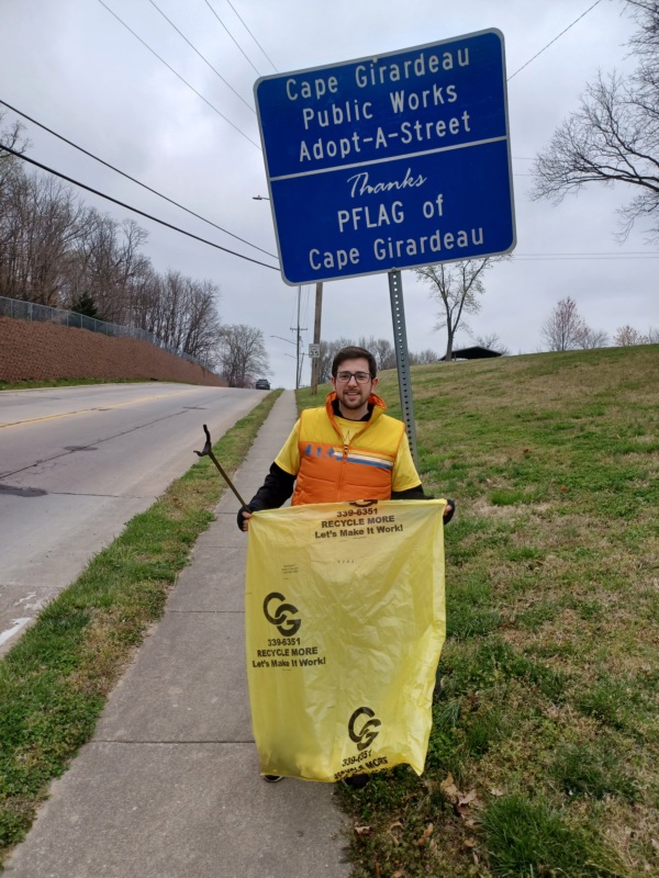 A member of Cape Girardeau PFLAG standing by the PFLAG Cape Girardeau Adopt a Road sign. The member has a trash pickup stick and a bag full of trash from the pickup day.