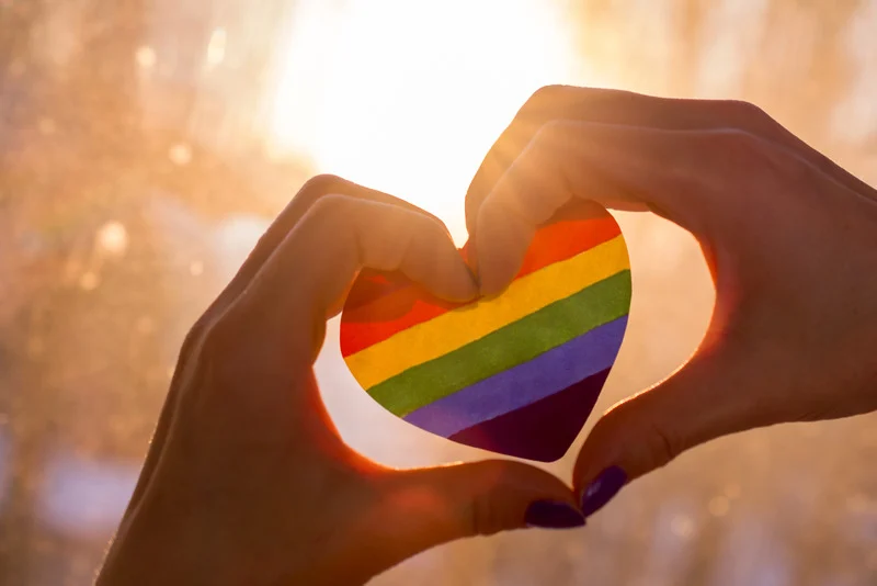 Image of someone's hand creating a heart with a pride heart in the middle. The sun is shining brightly in the background.